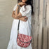 A woman wearing a white dress with a red and black floral sling bag over her shoulder.