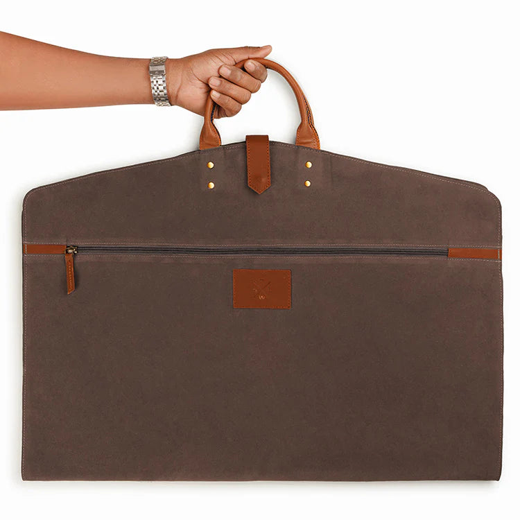 Brown garment bag with leather handles and trim, held by a hand on a white background