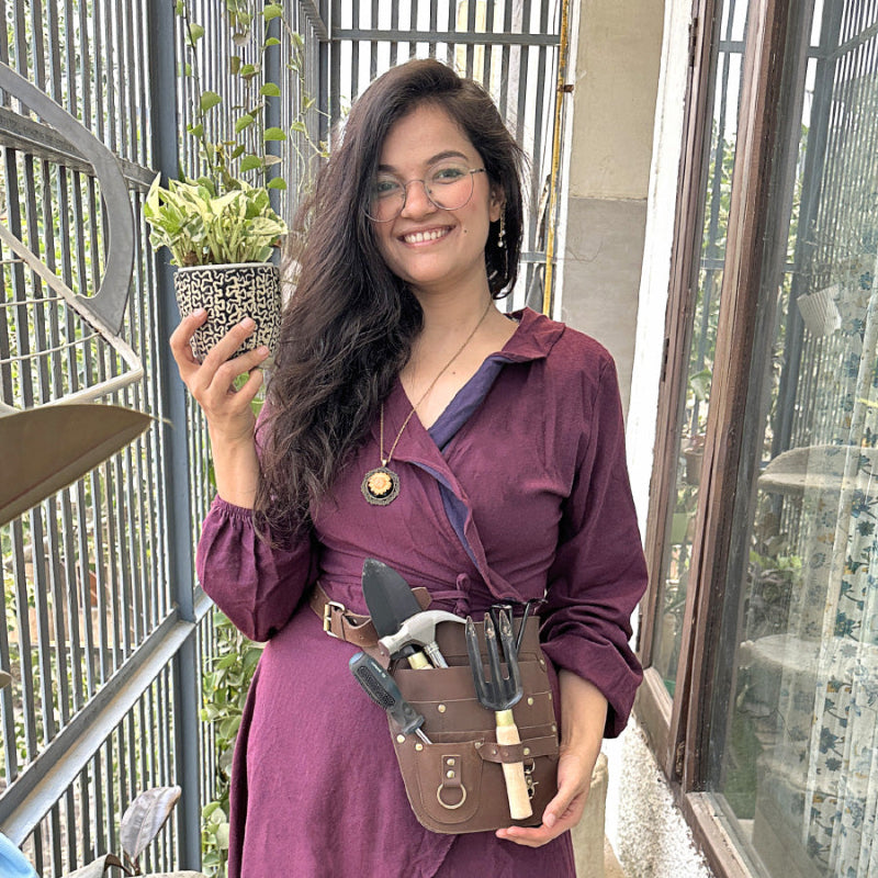 A woman in a purple dress holds a plant while wearing a leather tool belt around her waist.