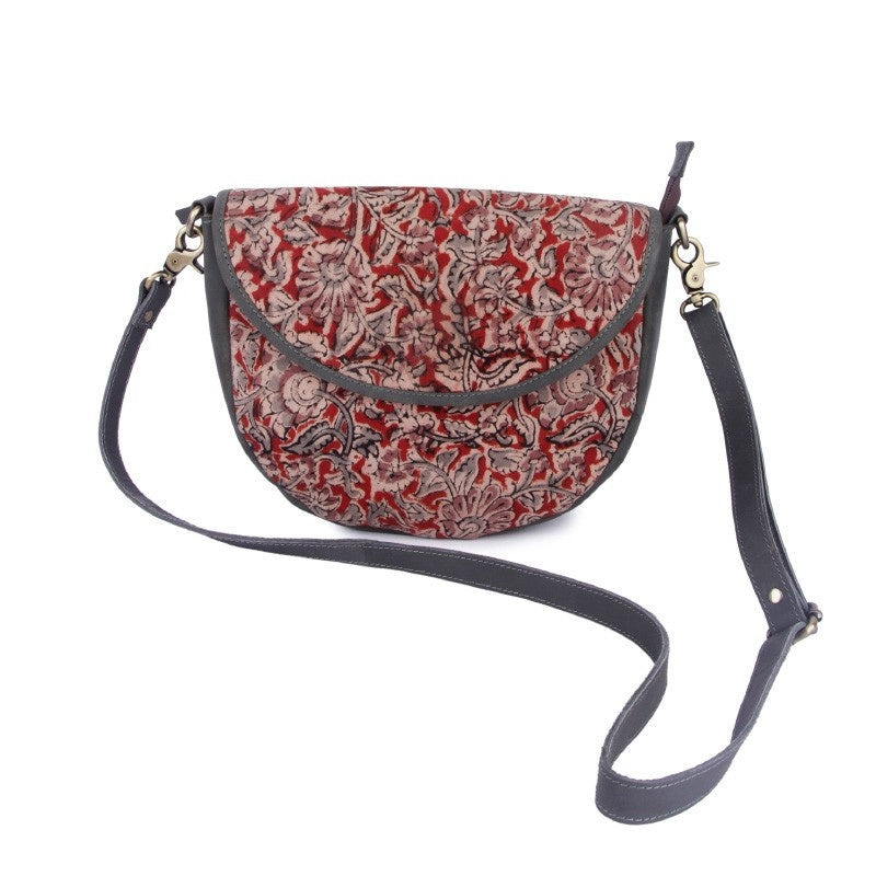 A sling bag with a red and black floral Kalamkari print and a black leather strap.