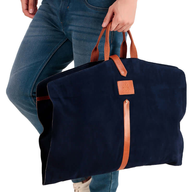 A black leather suit cover bag with brown trim and handles, laid out on a bed, displaying the interior with a suit hanging inside and compartments for storage.