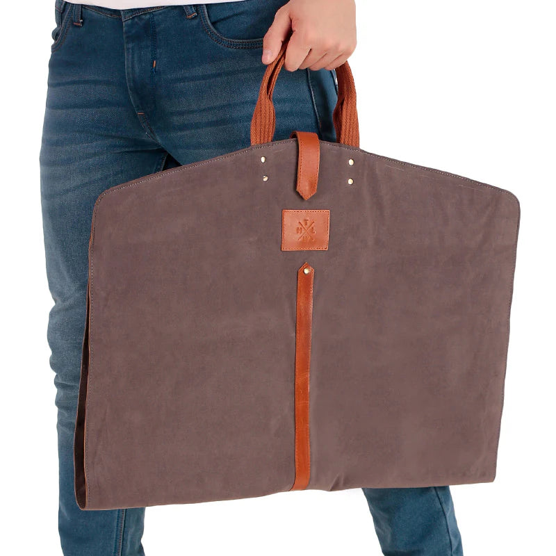 Brown garment bag with leather straps held by a person wearing blue jeans.