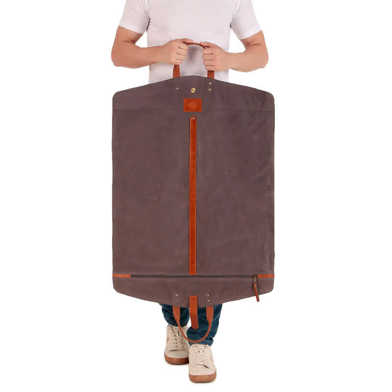 A black leather suit cover bag with brown trim and handles, laid out on a bed, displaying the interior with a suit hanging inside and compartments for storage.