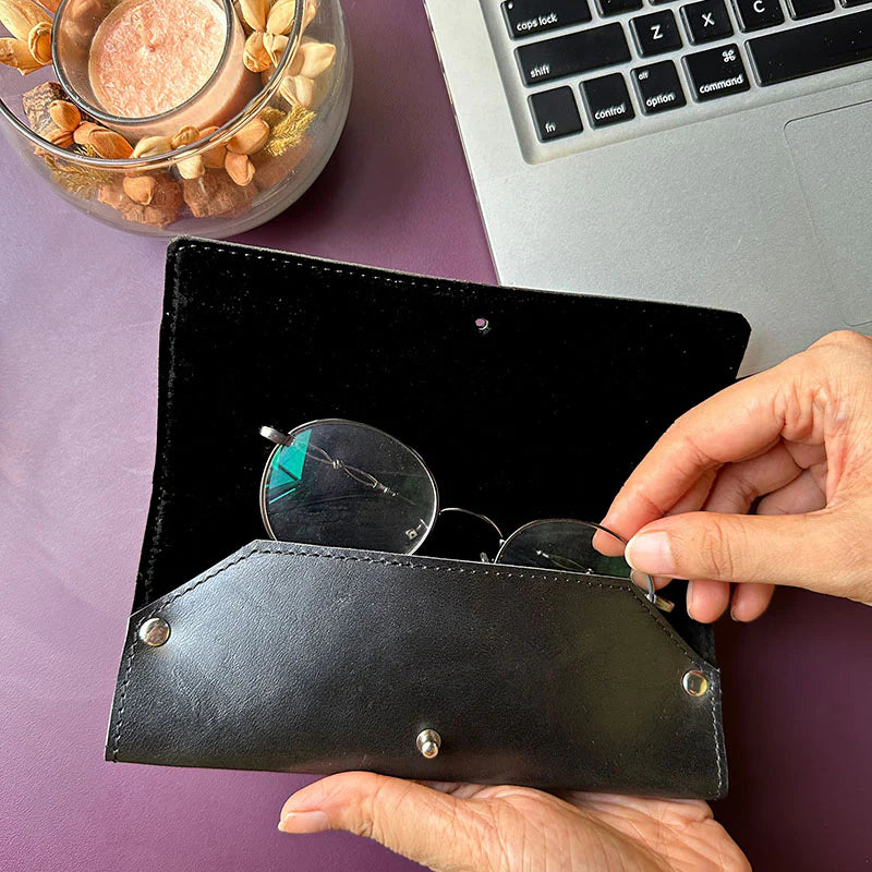 Person holding a black leather case with sunglasses on a purple surface next to a laptop.