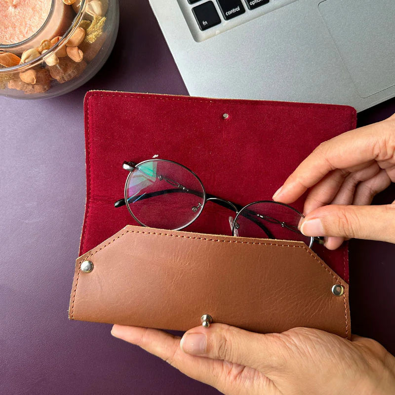 Person holding a brown leather glasses case with round eyeglasses on a red surface, with a laptop and container of snacks in the background.
