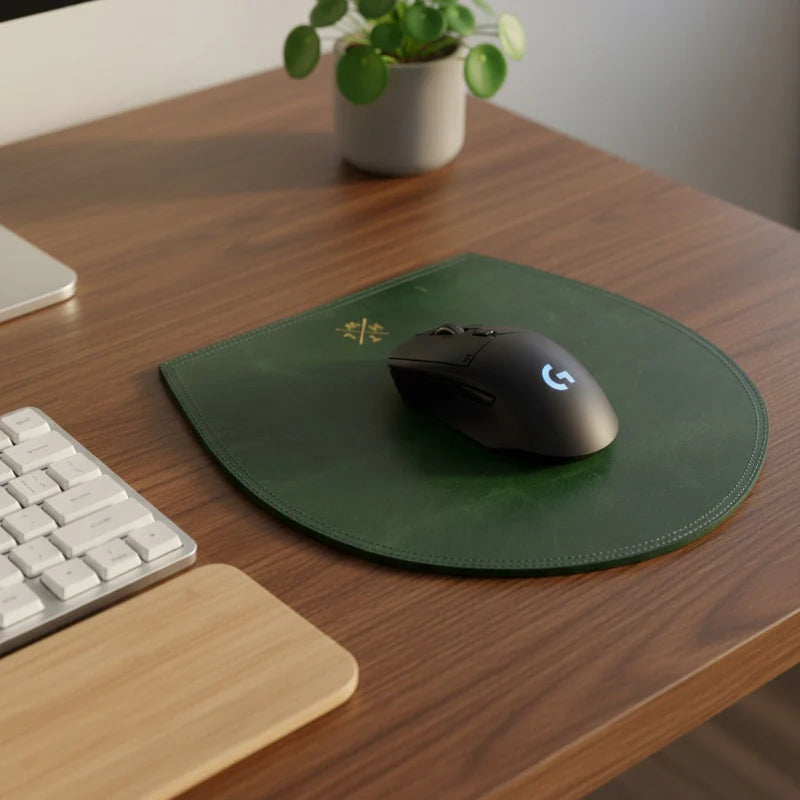 Black gaming mouse on a green mouse pad with a plant and keyboard in the background on a wooden desk.