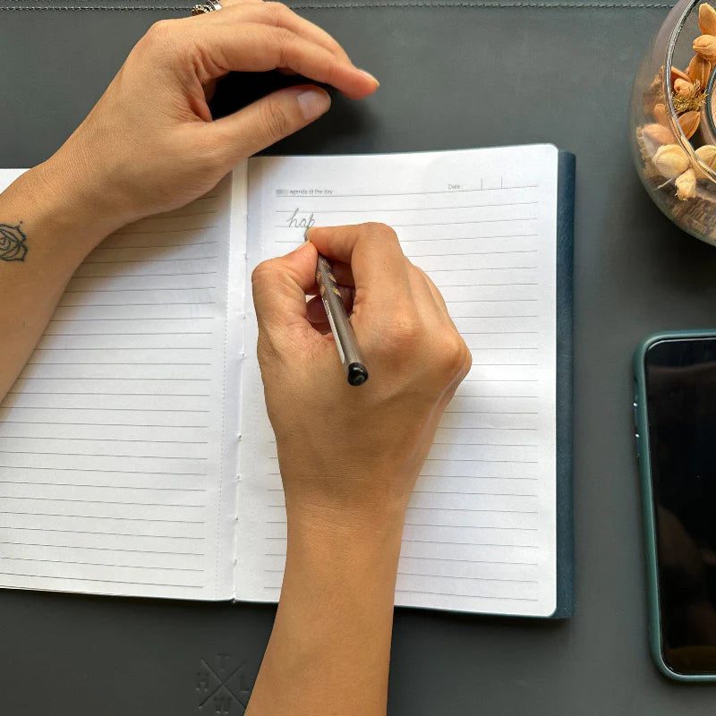 Person writing in a notebook with a pen on a desk, next to a smartphone and a small container.