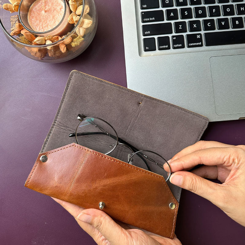 Person holding a brown leather eyeglass case with glasses, next to a laptop on a purple surface.