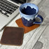 Blue mug on a brown leather coaster next to a laptop on a wooden surface