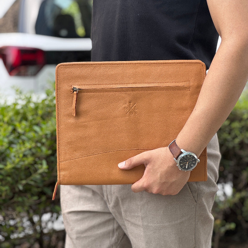 Person holding a brown leather clutch with a car and greenery in the background