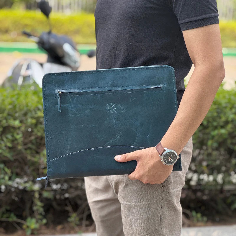 Person holding a dark blue leather clutch bag outdoors with greenery in the background