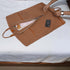 A brown leather suit carrier bag with handles and zippers, laid out on a bed next to a mobile phone.