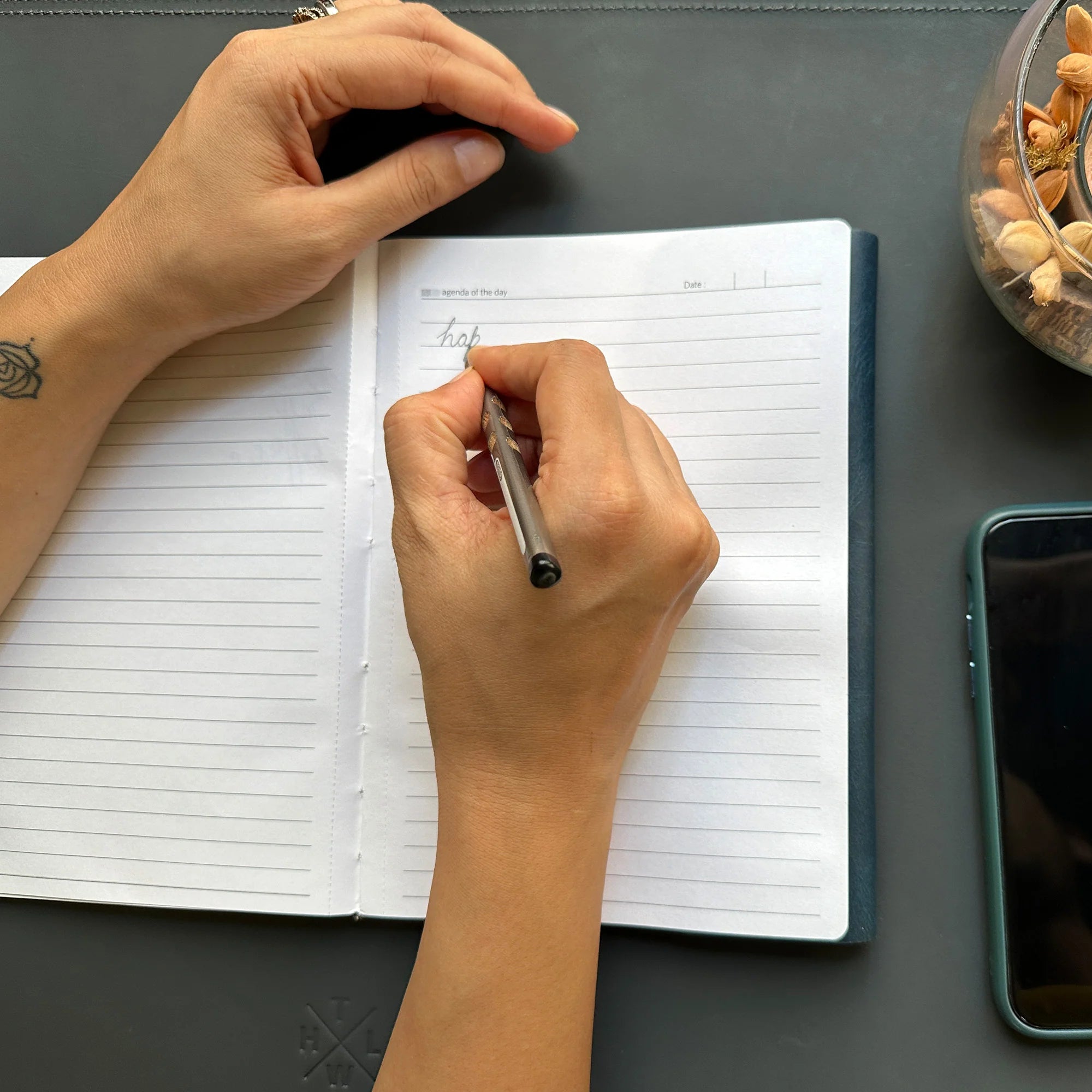 Person writing in a notebook with a pen on a desk, next to a smartphone and decorative items.