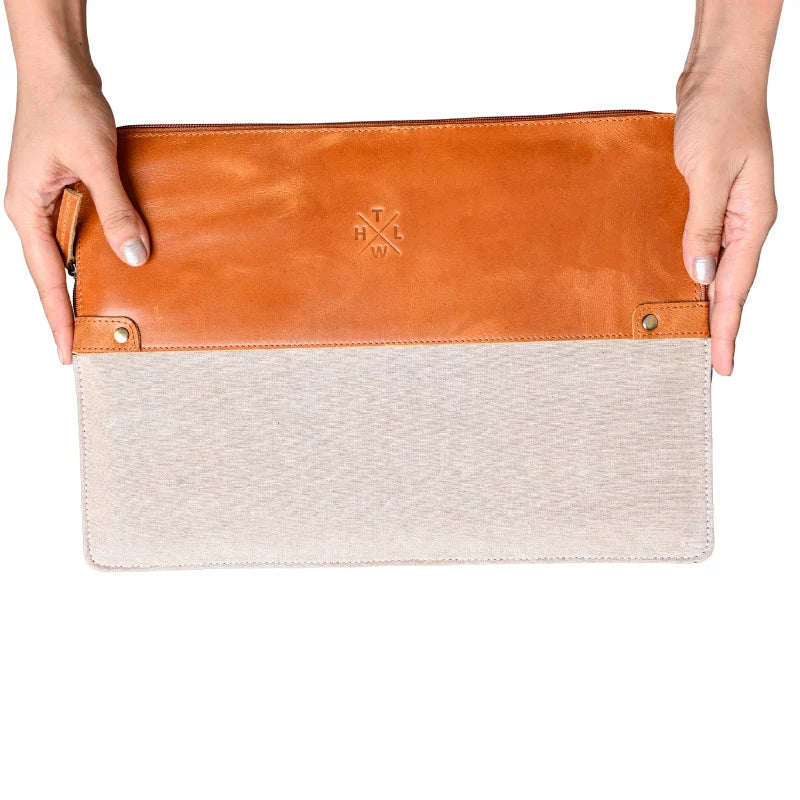 Two-toned clutch bag with brown leather top section and beige fabric bottom section held by hands on a white background.