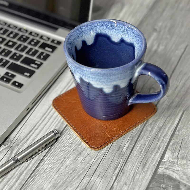 Blue ceramic mug on a brown coaster next to a laptop and pen on a wooden surface