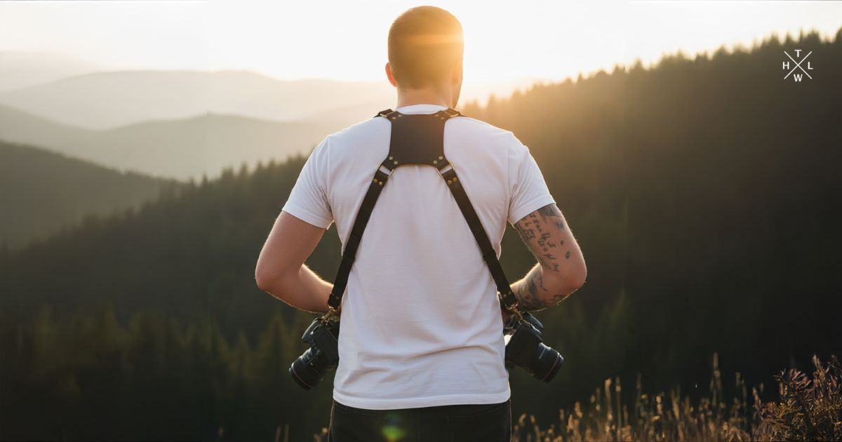 Photographer wearing a dual leather camera harness in the mountains during golden hour, holding two cameras with scenic background.