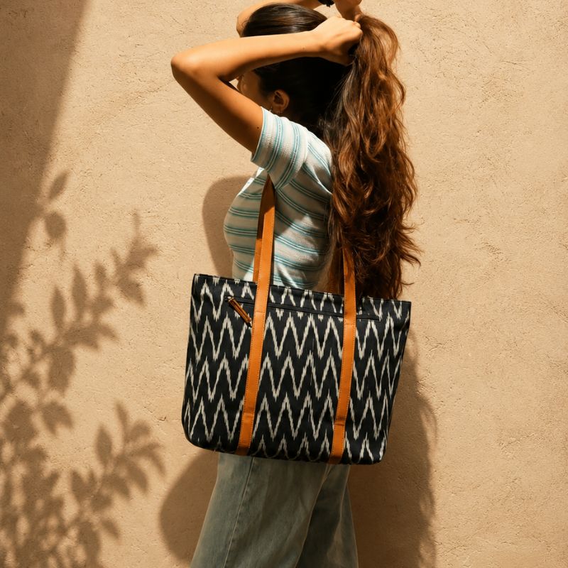 A woman with long brown hair stands against a textured beige wall, wearing a striped shirt and jeans while carrying a large tote bag. The bag has a black and white geometric zigzag pattern with tan shoulder straps.