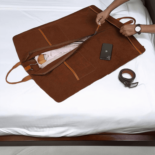 A brown leather garment bag with a soft velvet lining is laid out on a bed, with a person's hands adjusting its position. The bag has multiple compartments and a detachable shoulder strap, and it appears to be sturdy and high-quality.