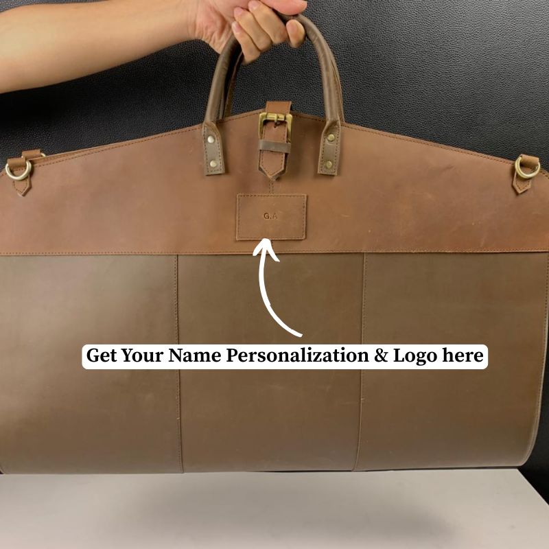 Brown leather garment bag  with a visible brand logo, held by a person against a dark background.