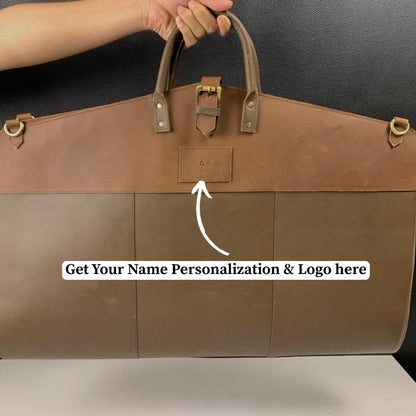 Brown leather garment bag  with a visible brand logo, held by a person against a dark background.
