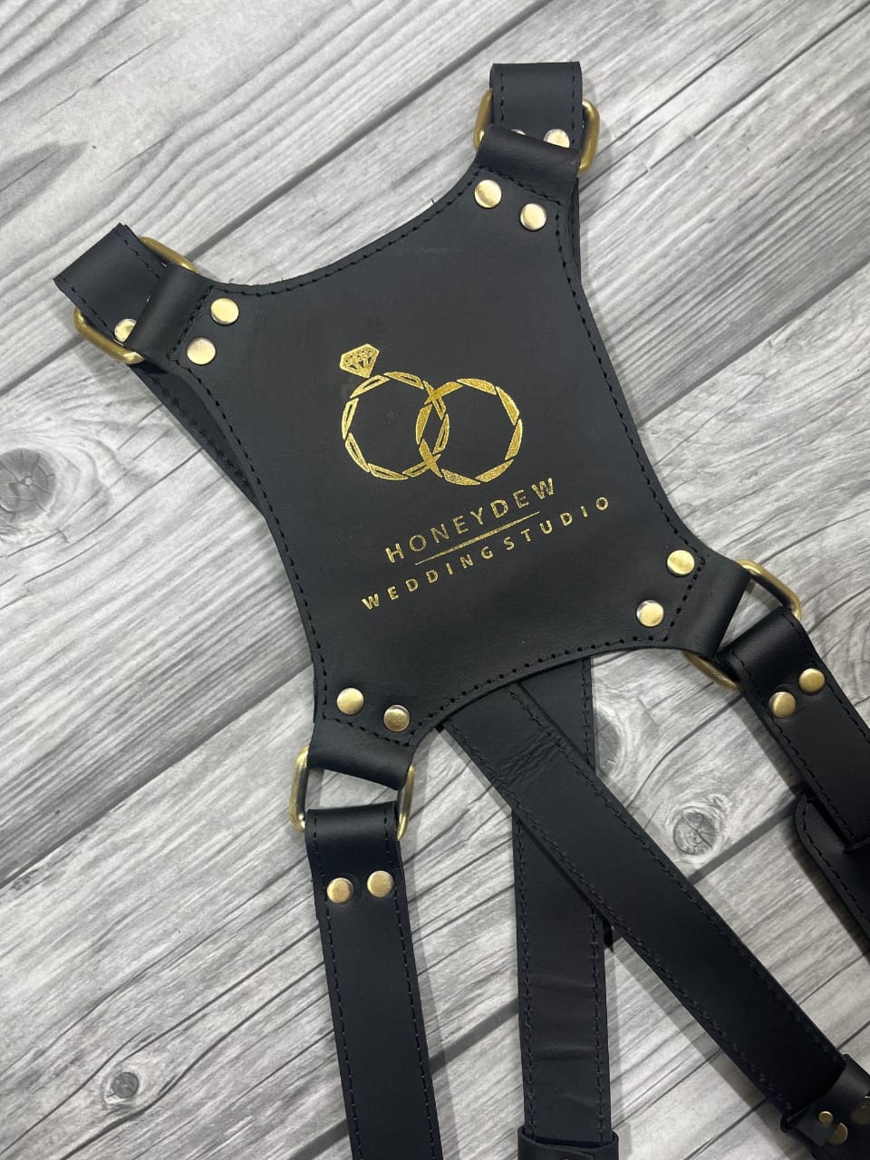 Black leather harness with gold accents on a wooden surface, featuring 'Honeypie Wedding Studio' branding.