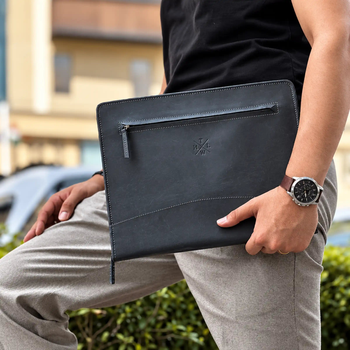 A person outdoors holding a black leather laptop sleeve or document pouch with a zippered pocket and an embossed logo. The person is wearing a black shirt, light gray trousers, and a brown leather-strap watch.