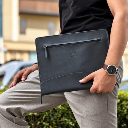 A person outdoors holding a black leather laptop sleeve or document pouch with a zippered pocket and an embossed logo. The person is wearing a black shirt, light gray trousers, and a brown leather-strap watch.