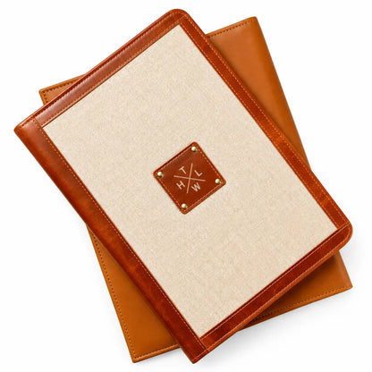 Beige canvas folder with brown leather trim, stitched edges, and a decorative leather logo patch