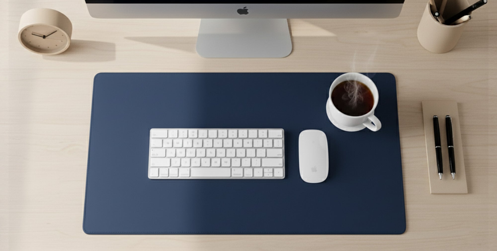 Blue desk mat on a light wooden desk with a white Apple keyboard, wireless mouse, and a steaming cup of coffee.