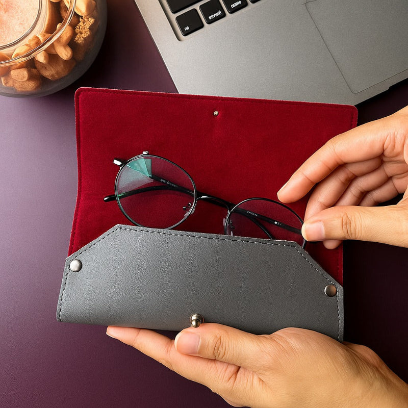 Person holding a gray eyeglass case with glasses on a red surface, with a laptop and snacks in the background.