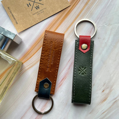 Two leather keychains on a light wooden surface with a perfume bottle and a card in the background.