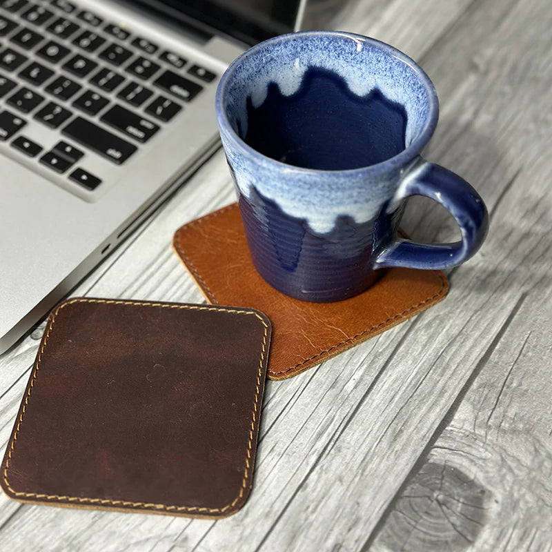 Blue mug on a brown leather coaster next to a laptop on a wooden surface