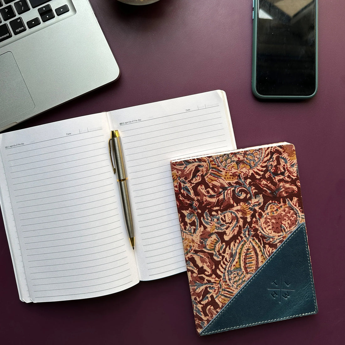 Open notebook with pen, patterned notebook, laptop, and phone on a purple surface