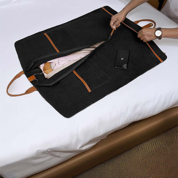 A black leather suit cover bag with brown trim and handles, laid out on a bed, displaying the interior with a suit hanging inside and compartments for storage.