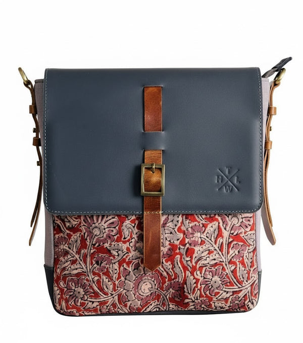 A front-view product shot of a rectangular messenger bag featuring a dark grey leather top flap with a vertical brown leather buckle strap. The bottom half of the bag is made of a fabric with an intricate red and cream floral pattern. The bag is set against a solid white background.
