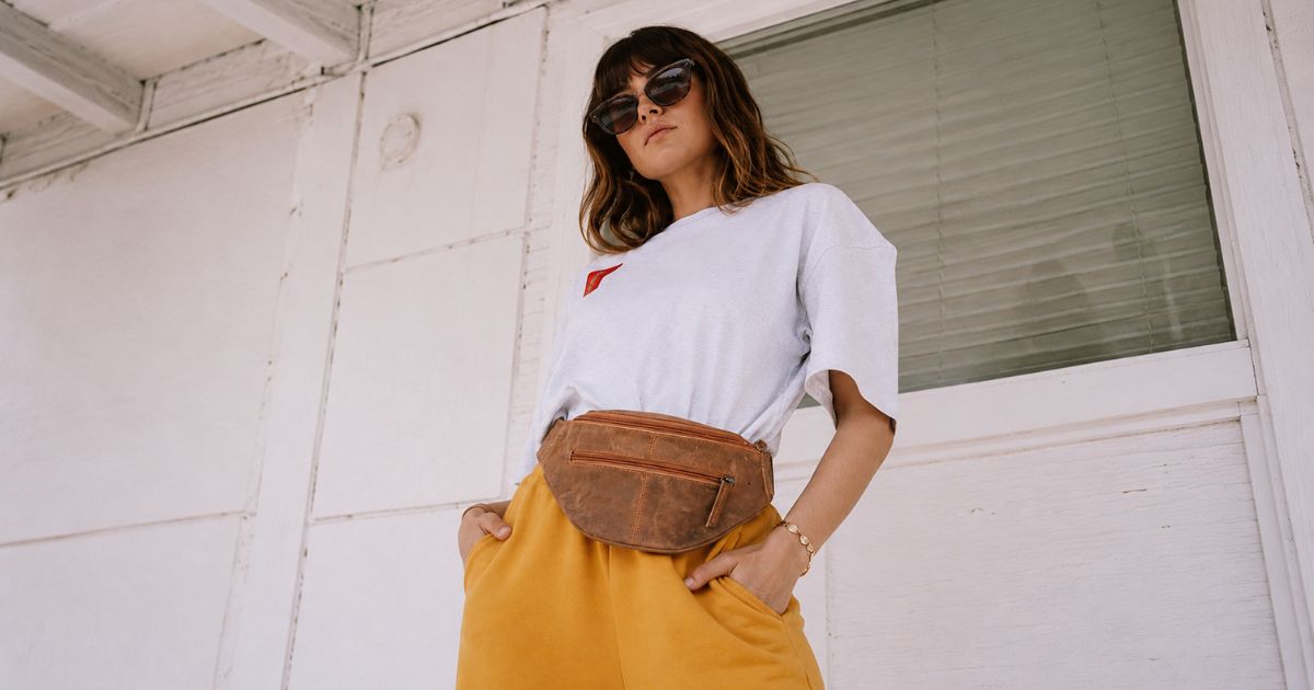 A woman wearing a white t-shirt, yellow skirt, and brown leather fanny pack, standing against a white wooden wall.