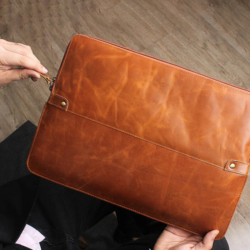 Brown leather clutch bag held by a person on a wooden floor.